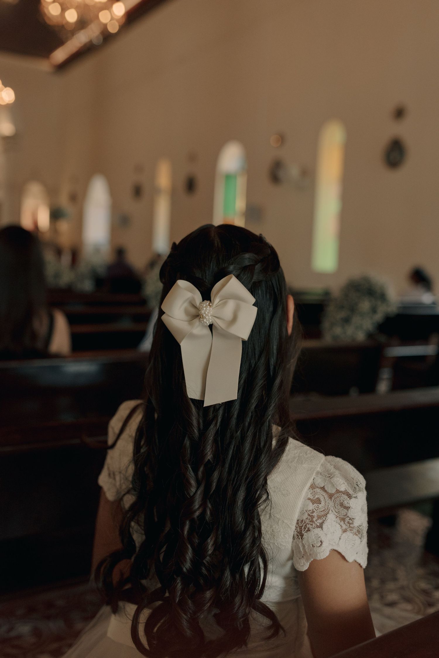 Long dark curled hair adorned with a white bow shown from behind in a church setting.