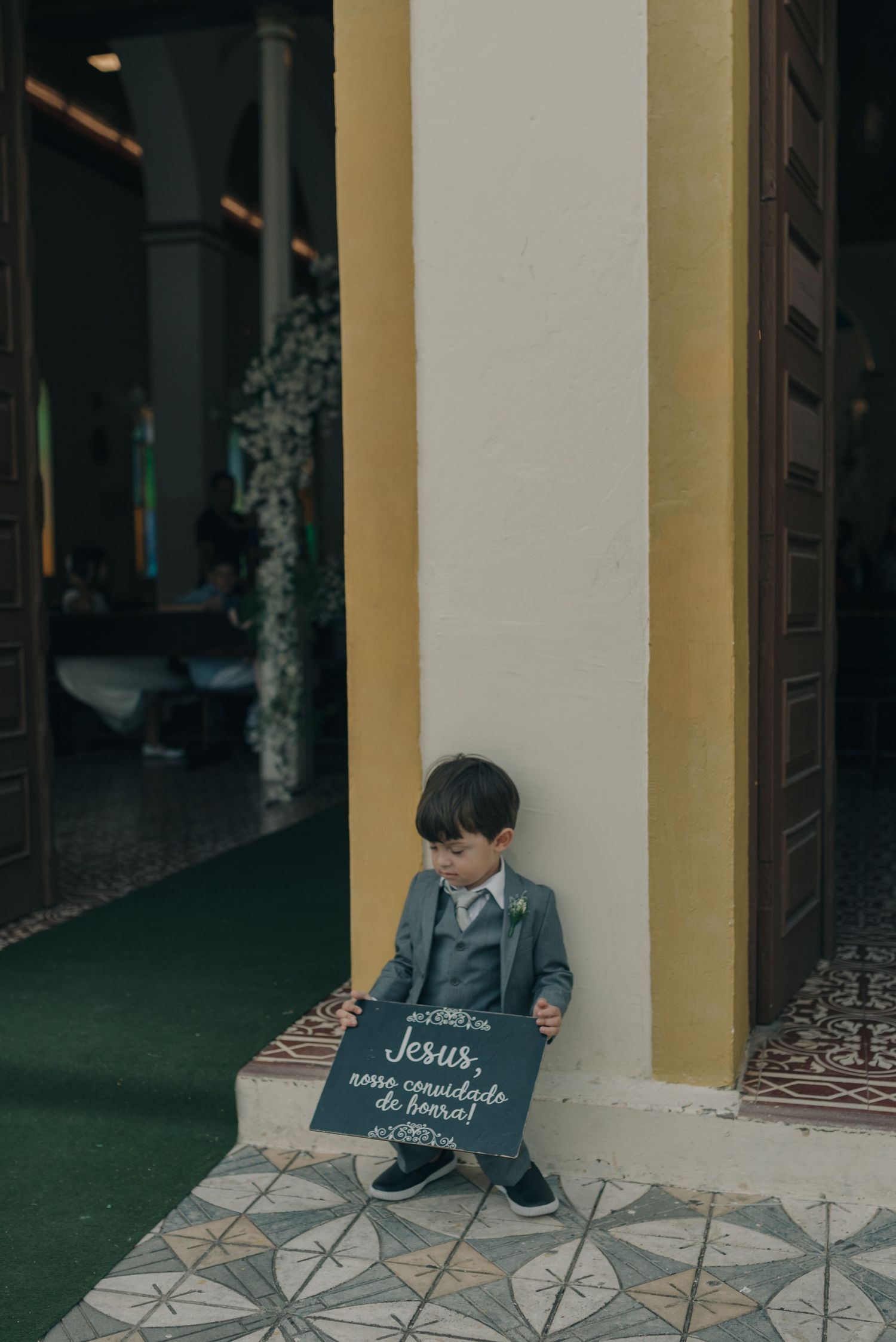 Small attendant in grey suit holds a wedding sign while standing against a yellow pillar.