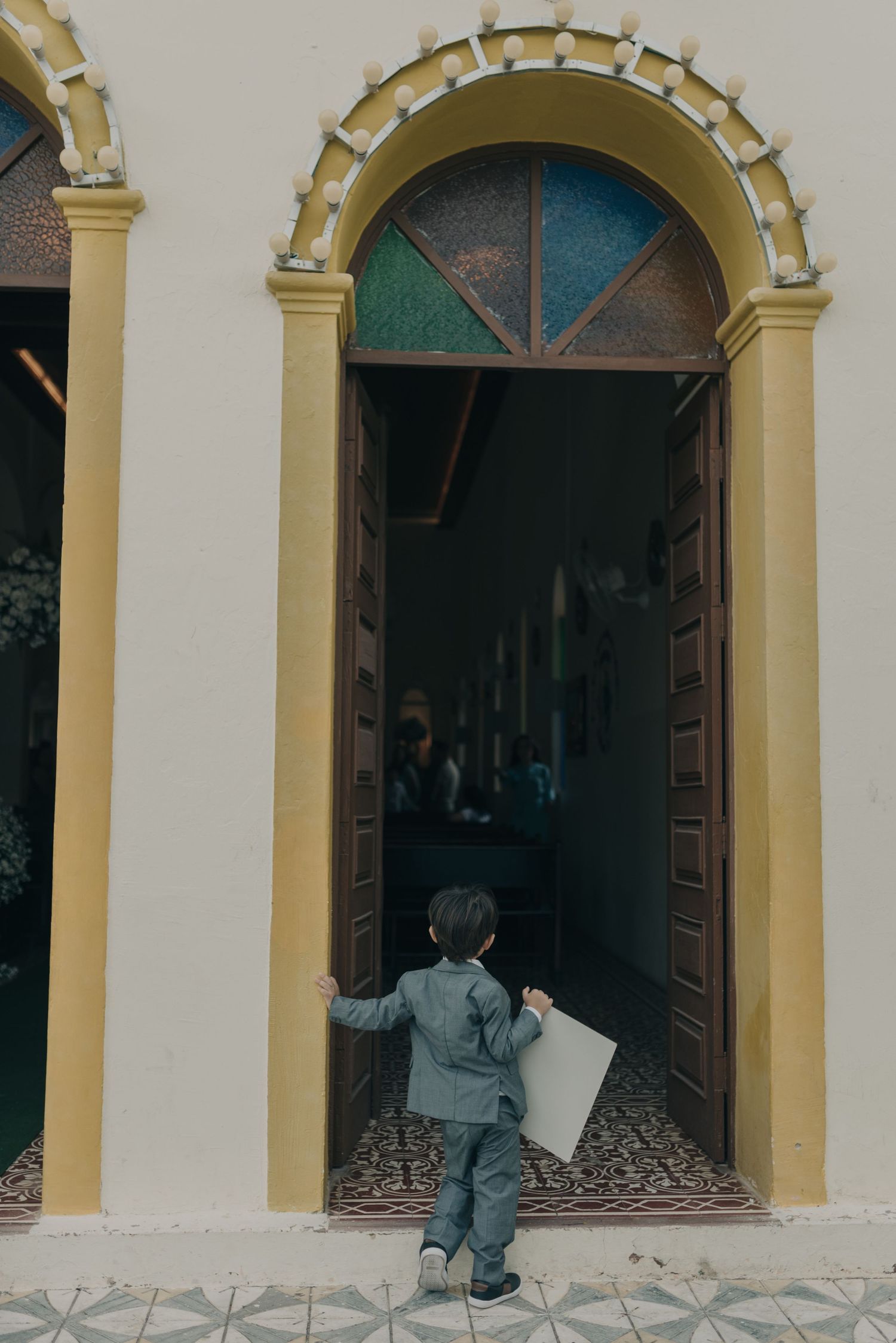 Young wedding attendant in grey suit approaches ornate church doorway with arched architecture.