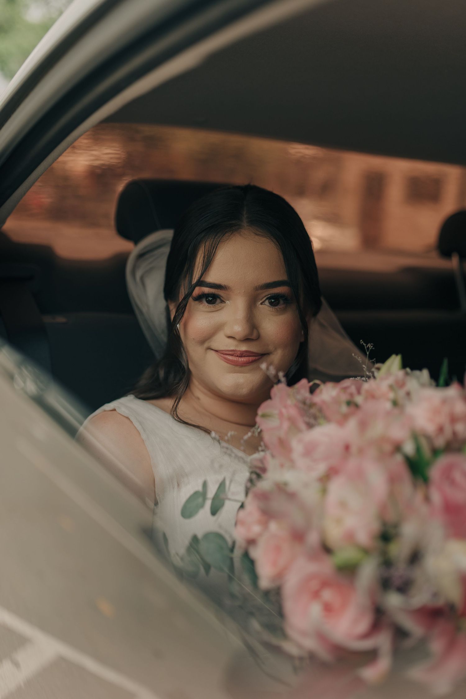 Person in bridal attire sits in car with pink rose bouquet visible through window.