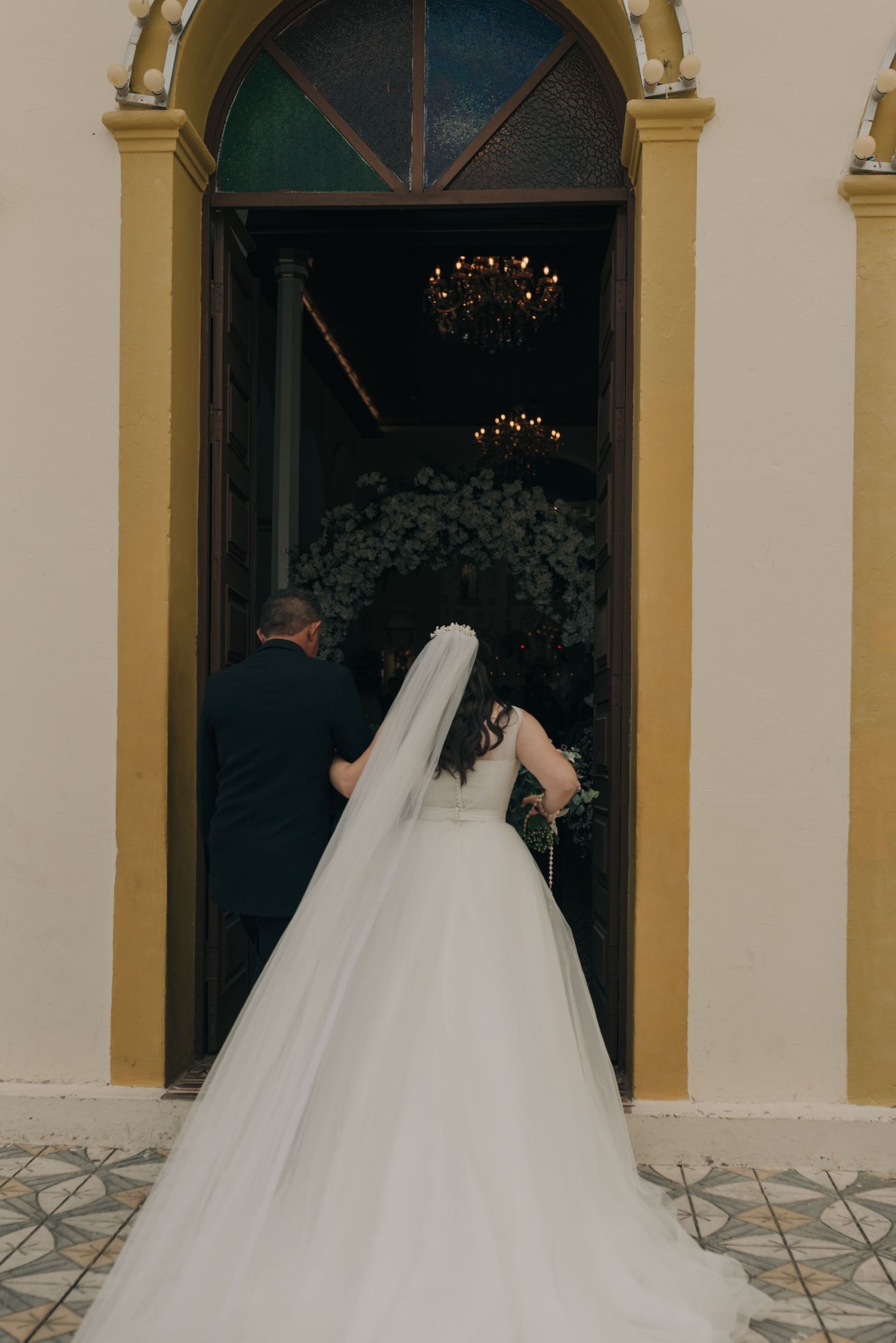 Back view of a bride in flowing white gown entering through an ornate yellow-framed doorway.