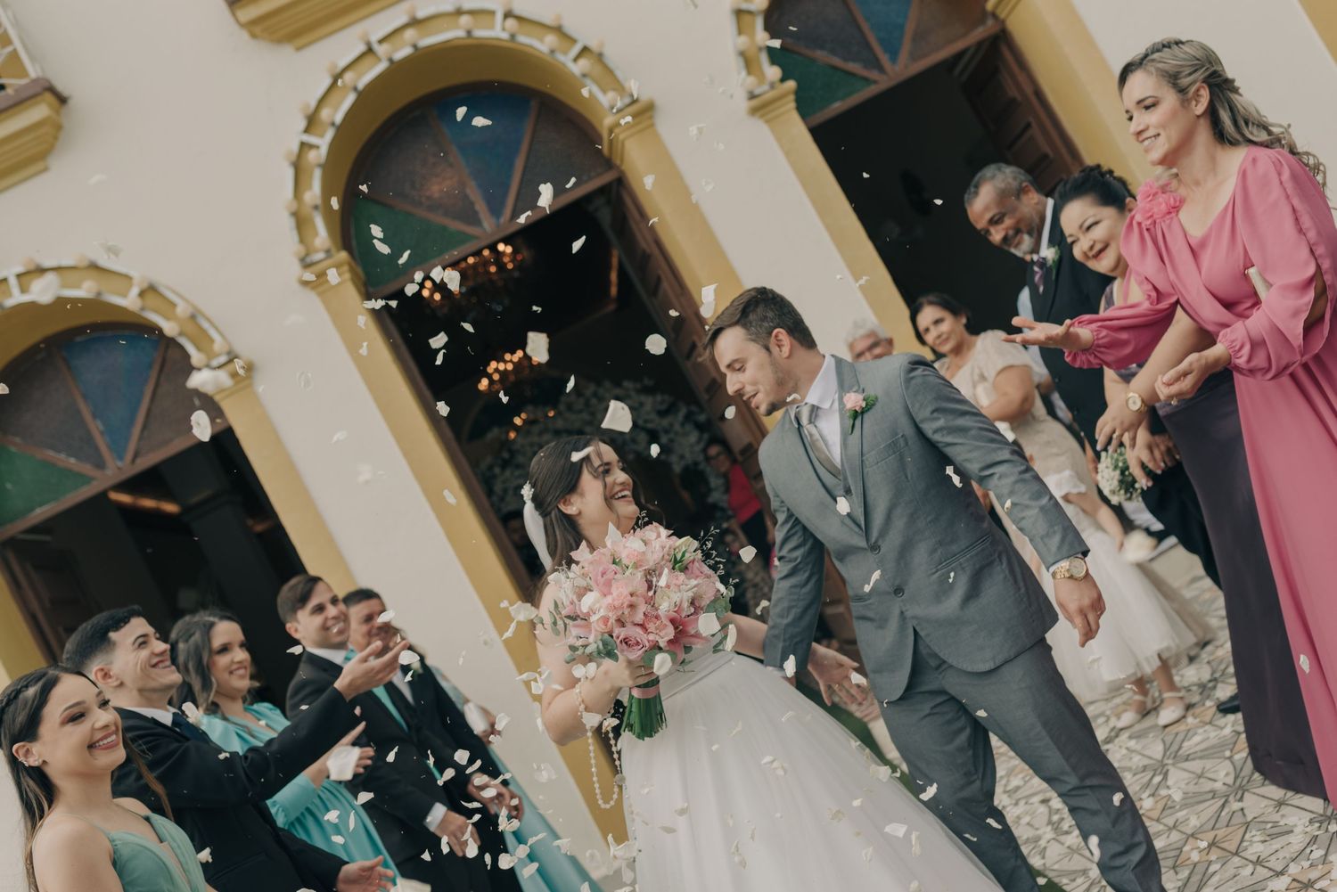 Wedding party celebrates with confetti outside a historic building with ornate arched windows.
