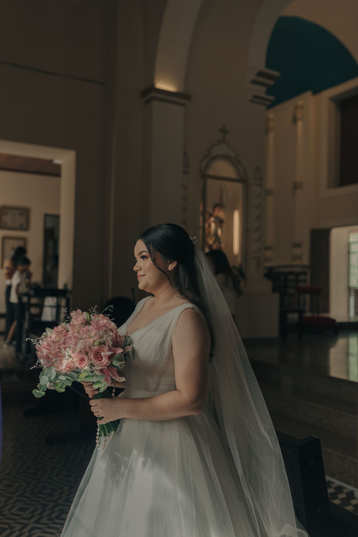 A bride in a white ballgown holds a pink bouquet in an elegant church interior.