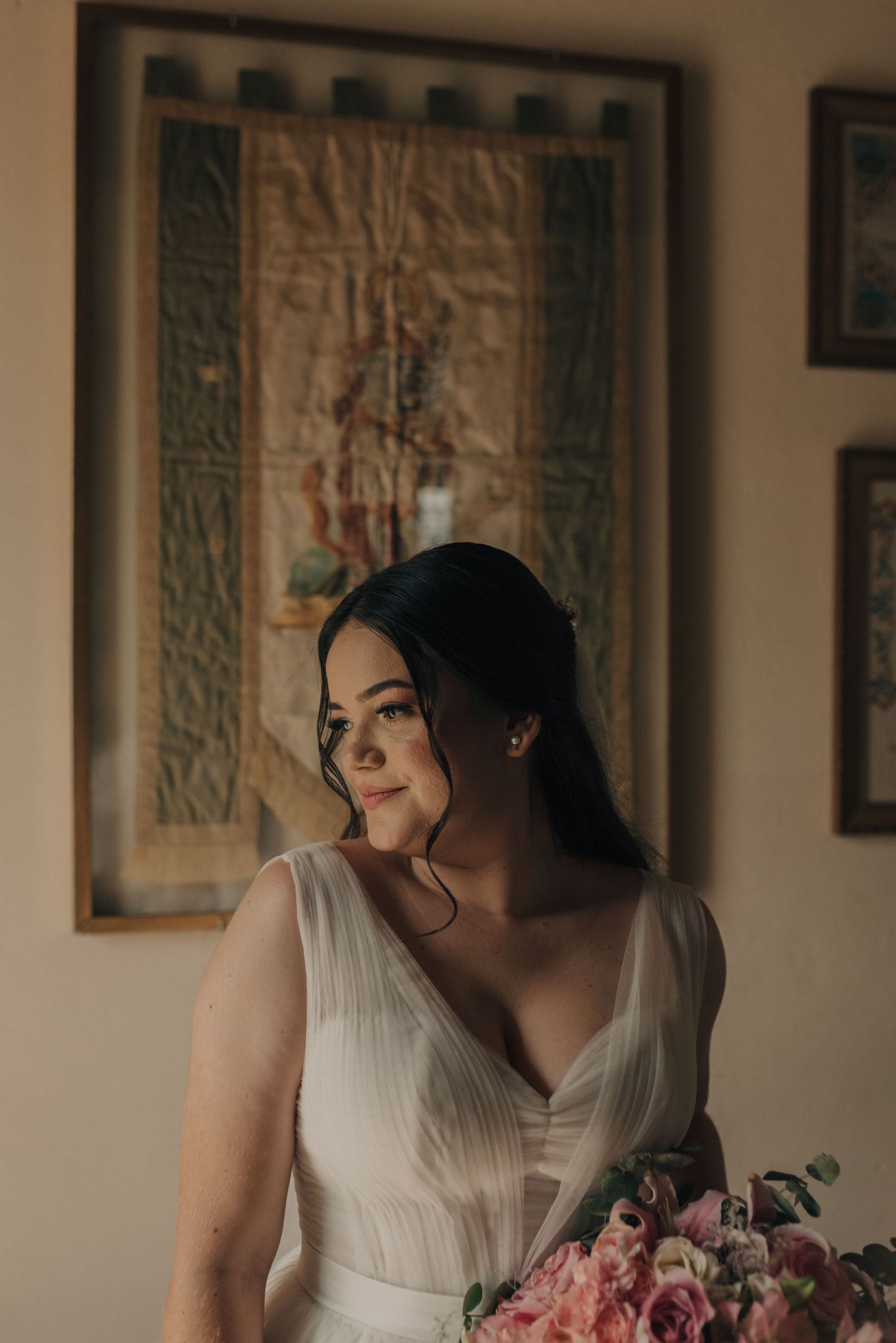 A bride in white holds a pink bouquet while posing against decorative wall hangings.