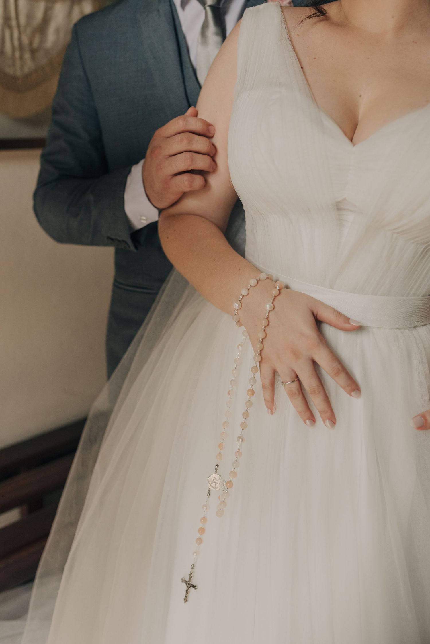 Close-up detail of a rosary bracelet and wedding dress with groom in background.
