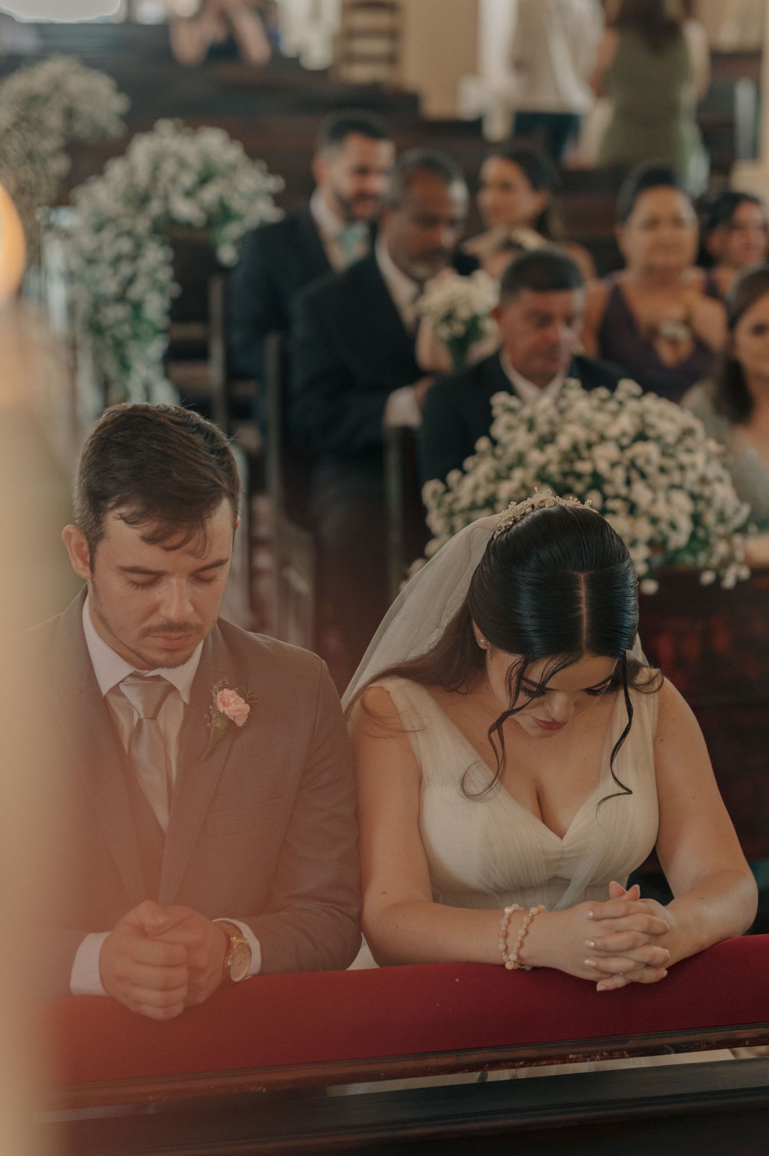 A couple bows their heads in prayer during a church wedding ceremony surrounded by white floral arrangements.