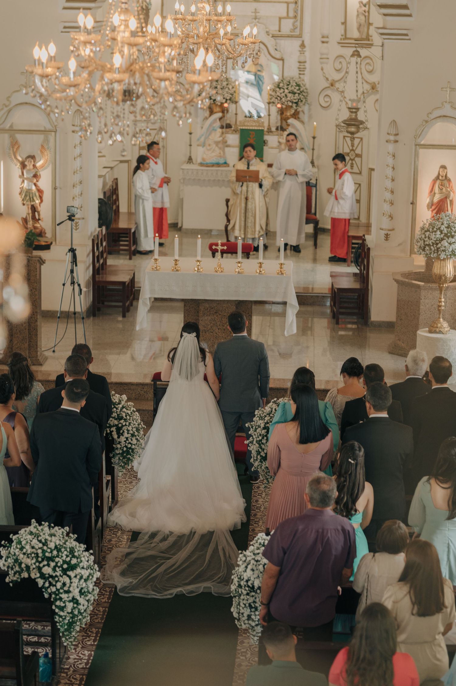 Wide shot of a Catholic wedding ceremony with altar servers, flowers, and chandelier in an ornate church.