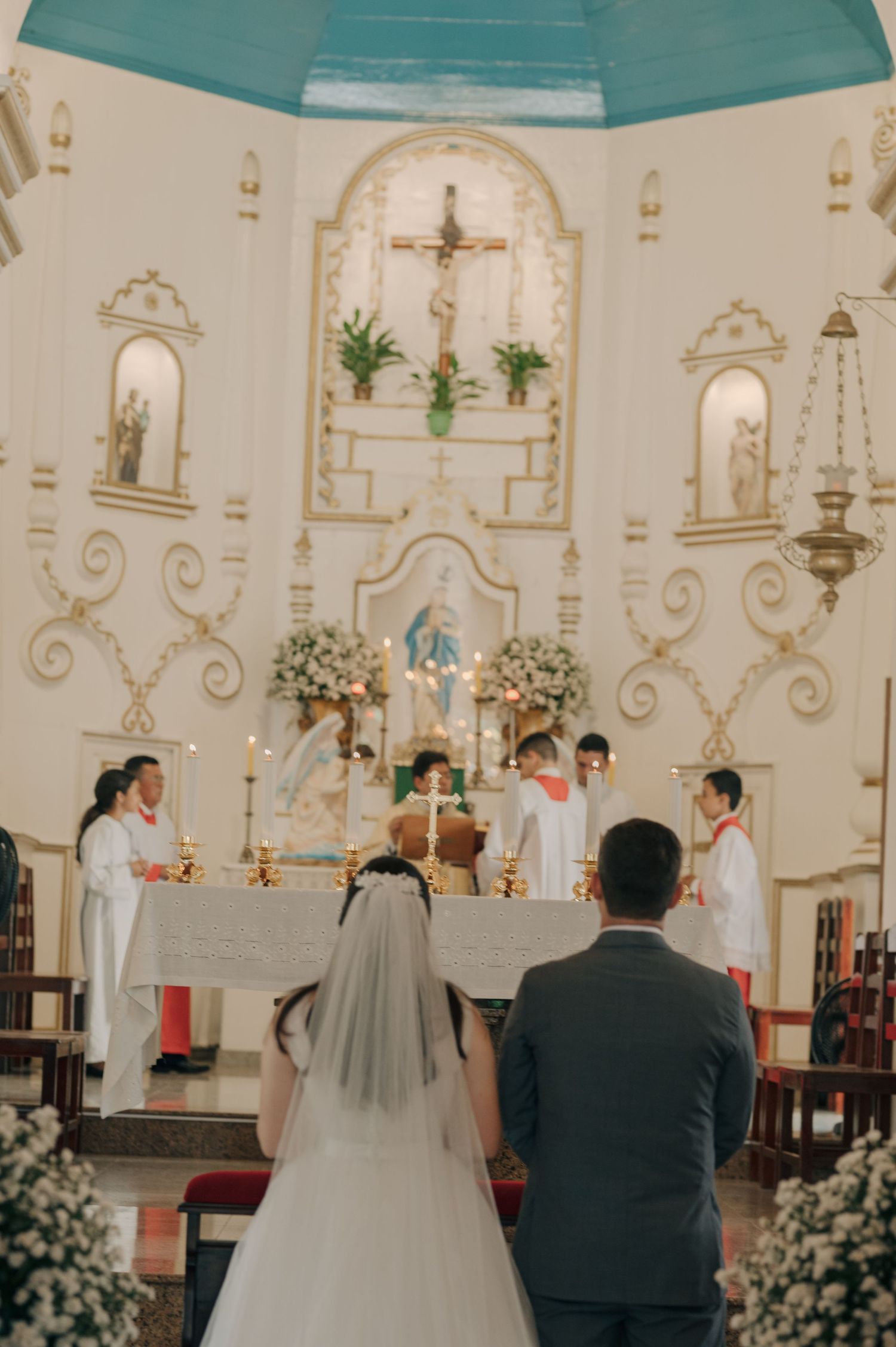 A wedding ceremony takes place at an ornate Catholic church altar decorated with flowers and crosses.