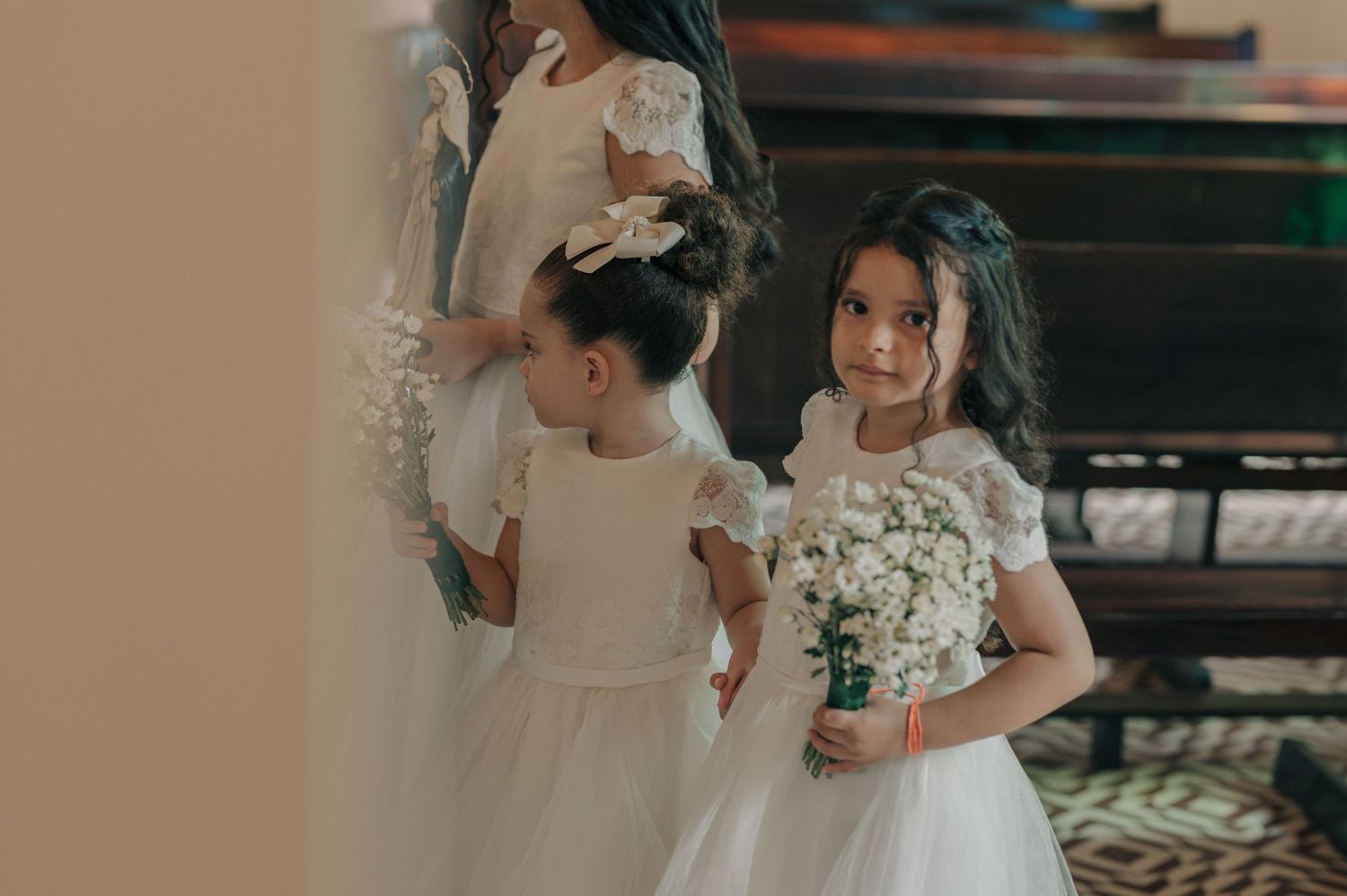 Young attendants in white dresses hold bouquets while participating in a wedding ceremony.