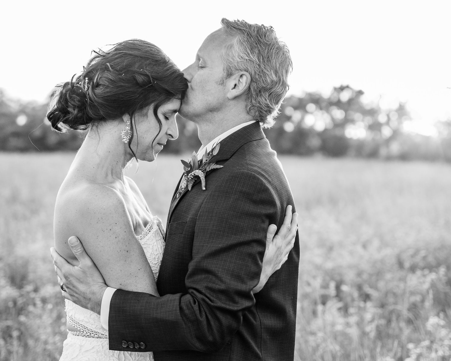 Husband kissing bride on her forehead in a field at sunset
