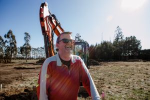 Worker in safety gear standing by construction equipment on dusty work site.