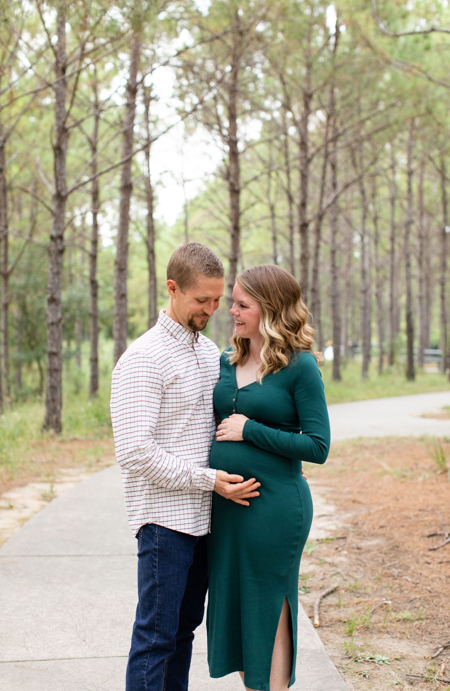Romantic maternity photo shoot featuring a couple on a pine tree-lined path with the woman in an elegant green dress.