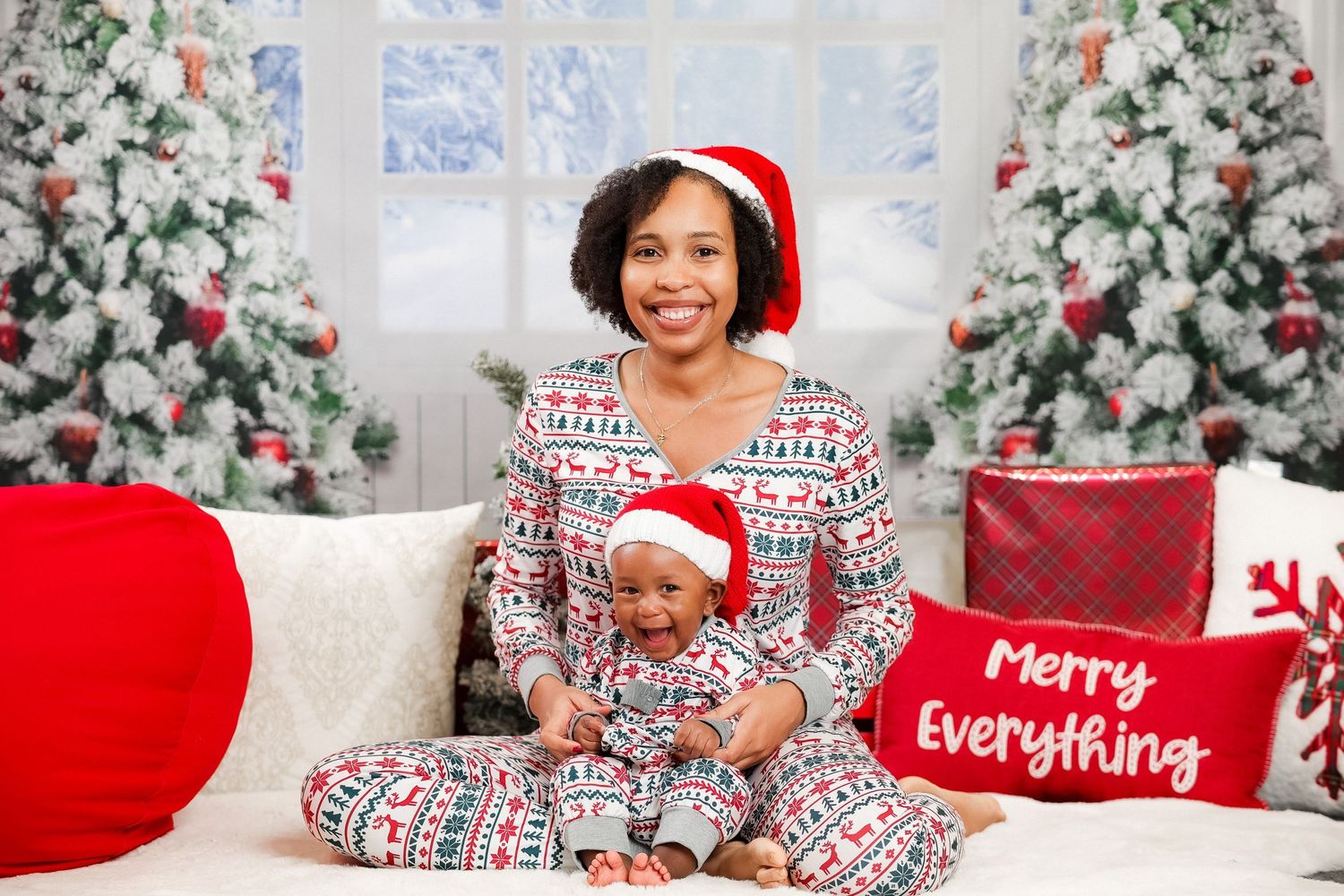 Mother and baby in matching Christmas pajamas sitting together near decorated holiday trees.