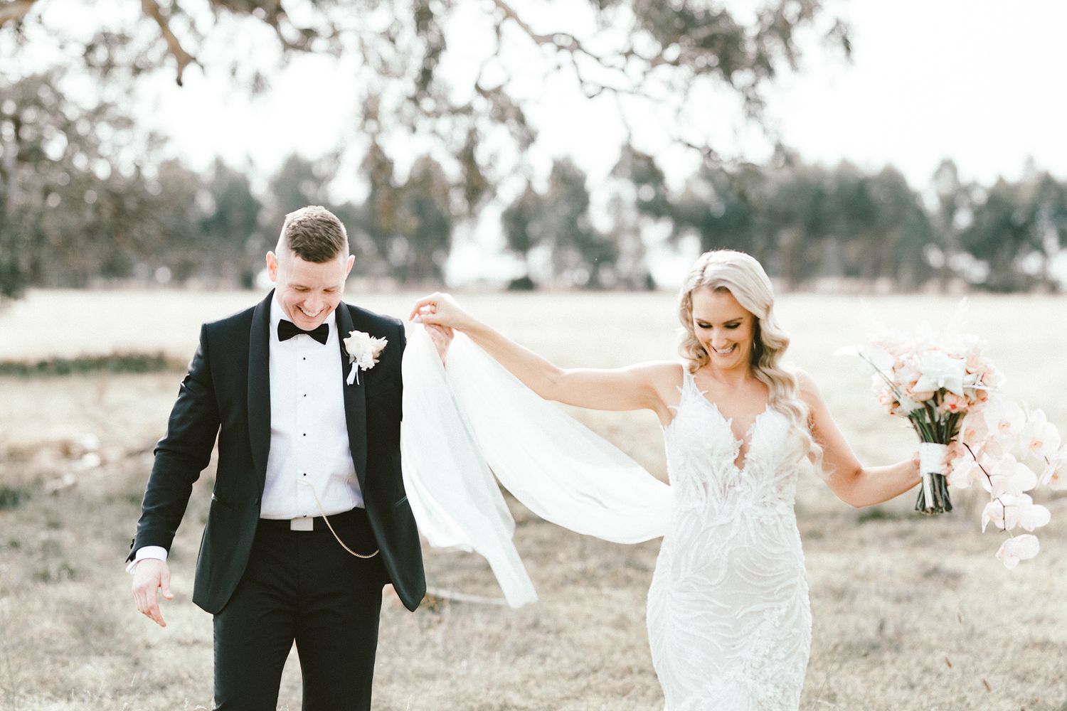 A romantic wedding portrait shows a couple walking through a rustic outdoor field.