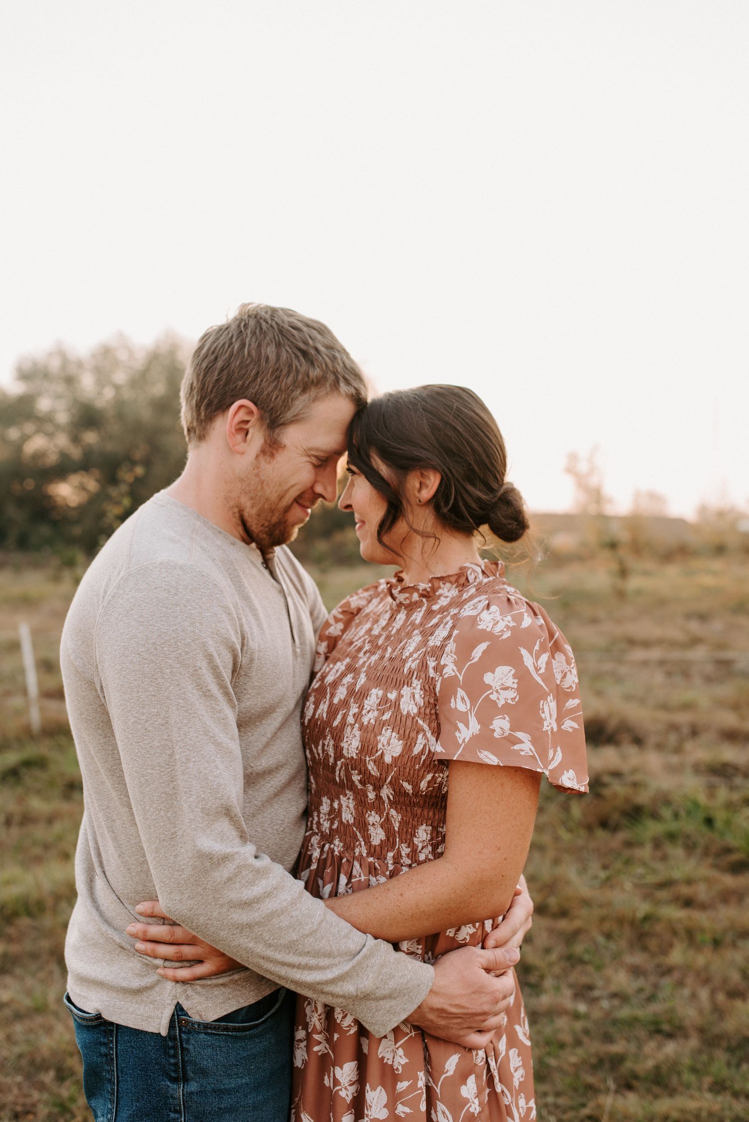 Intimate couple embraces in a field during golden hour wearing floral dress and beige sweater.