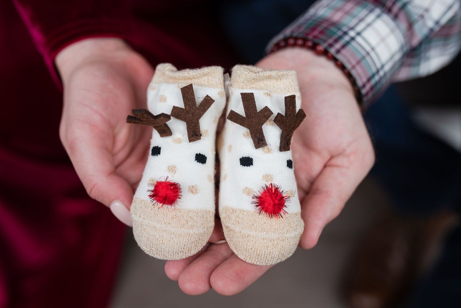 Pair of white reindeer-themed baby booties with red noses held in someone's hands.
