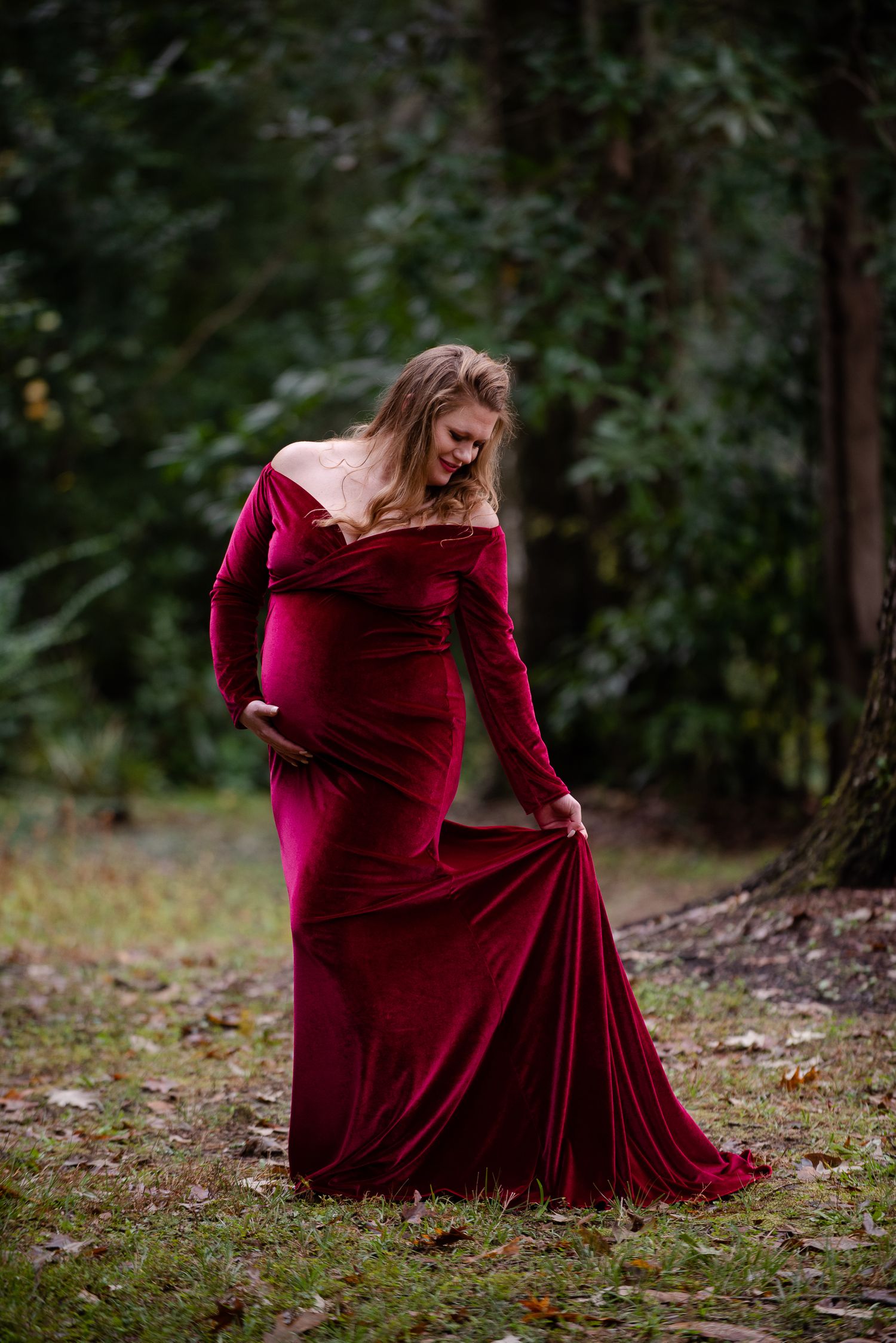 Person in a flowing burgundy velvet dress poses dramatically in a wooded setting.