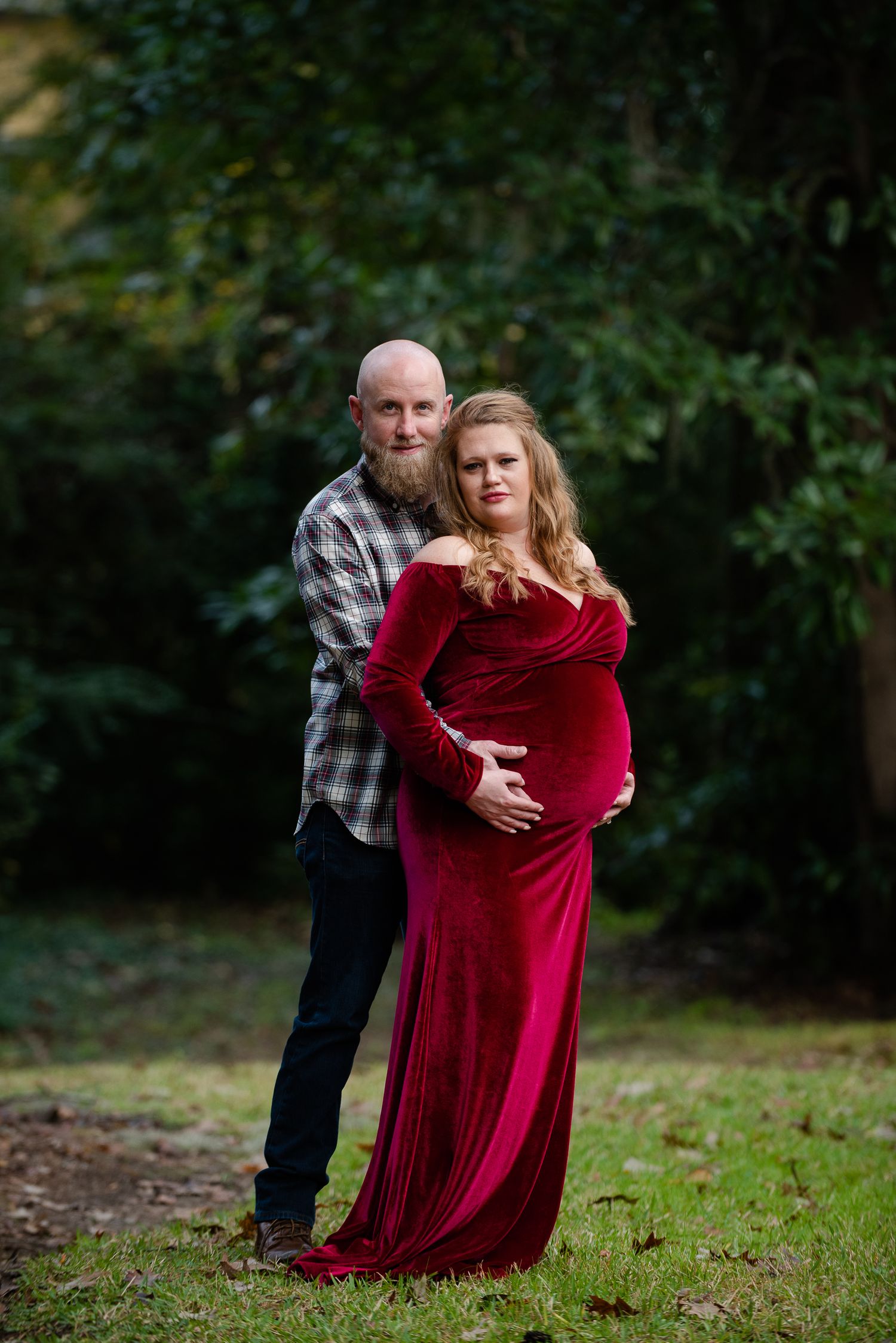 Couple embracing in an outdoor maternity photoshoot wearing red velvet dress and plaid shirt.