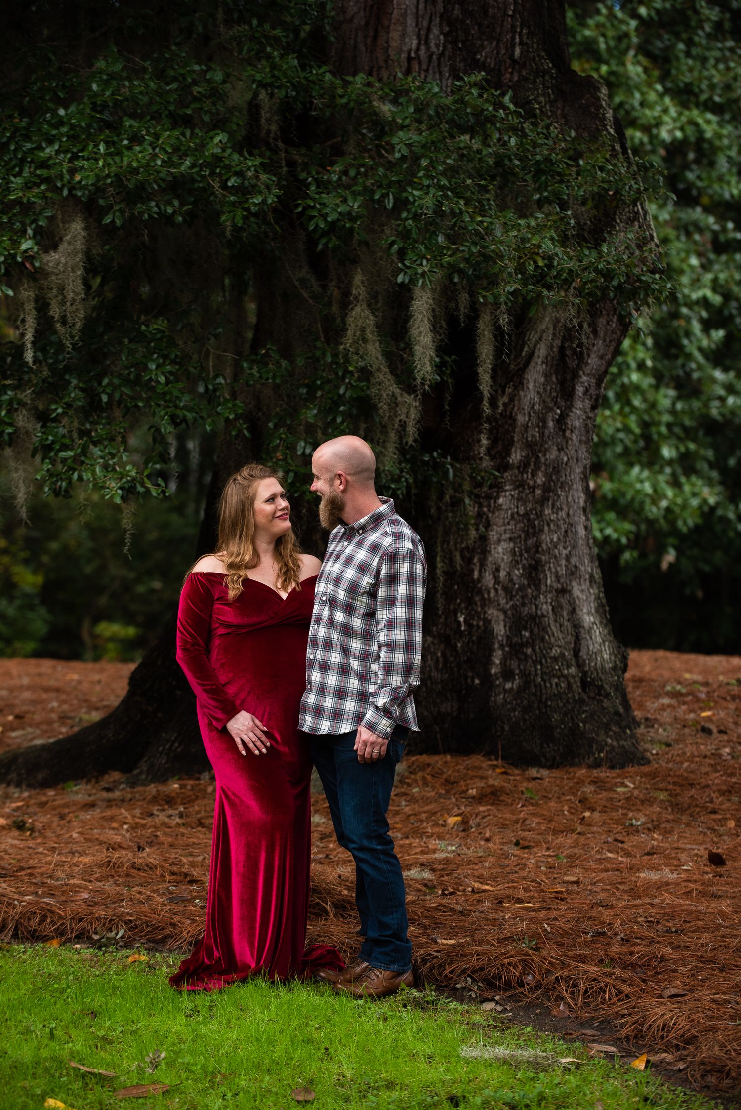 Couple standing by mossy tree during outdoor maternity photoshoot.