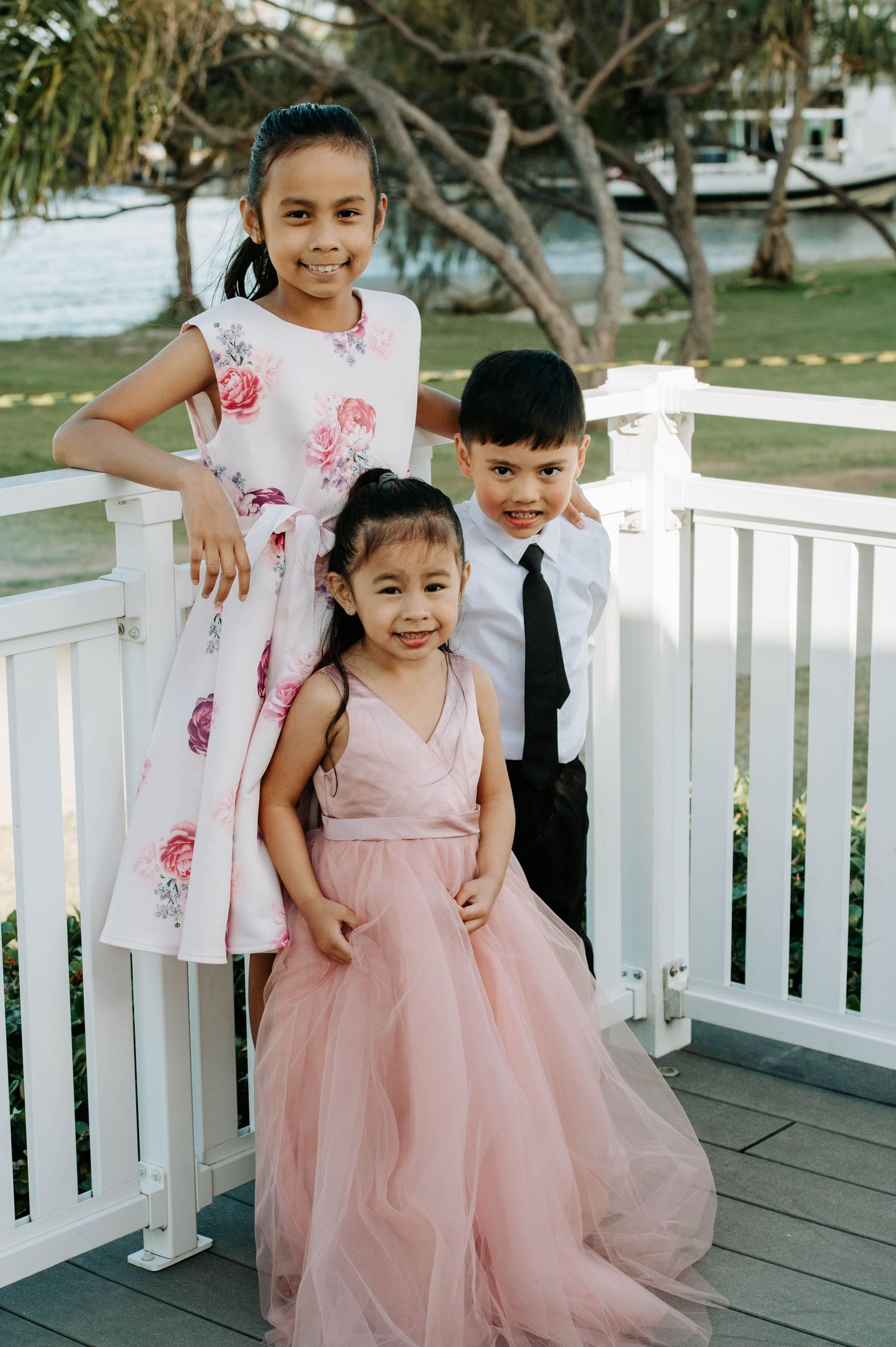 Three children in formal attire pose together on a white deck with palm trees in the background.