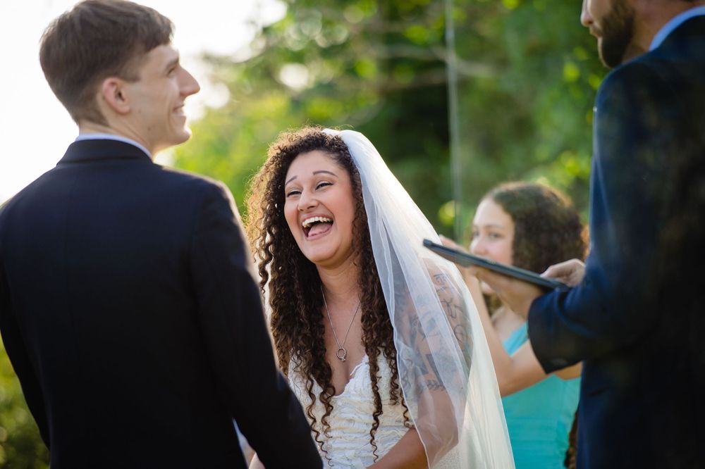 Happy bride in a white dress and veil shares joyful laughter during an outdoor wedding ceremony.