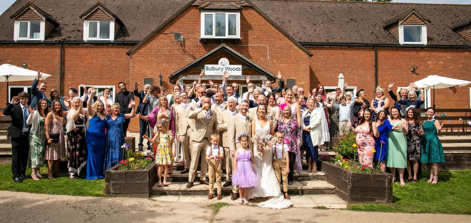 Large group of wedding guests gathered outside a rustic brick building for a celebratory photo.