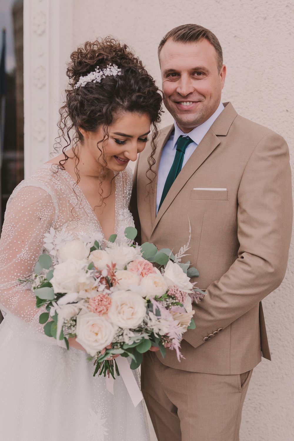 Newlyweds pose together with bride holding romantic pastel bouquet against neutral wall.