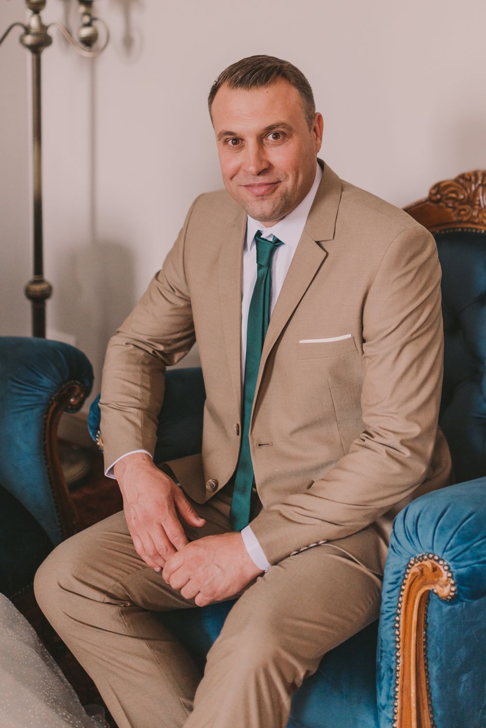 Groom in tan suit with teal tie sits on vintage blue velvet couch for formal portrait.
