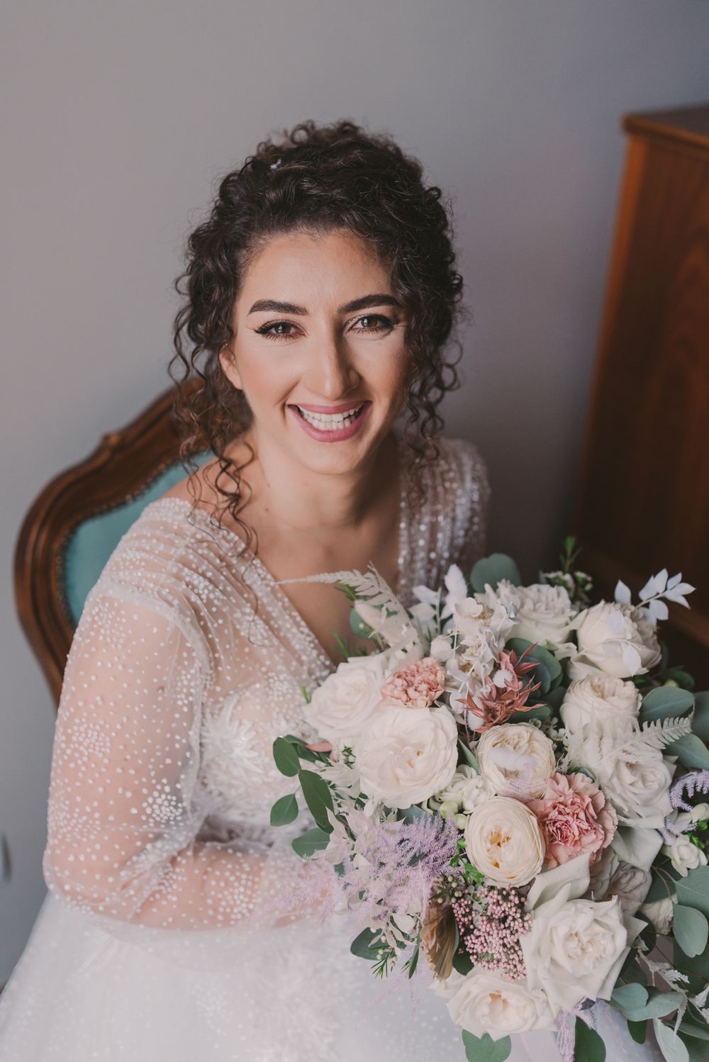 Bride holds romantic pastel bouquet featuring roses and greenery against white lace dress.