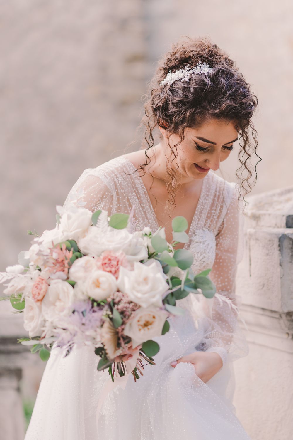 Bride in detailed lace gown admires her soft pink and white wedding bouquet.