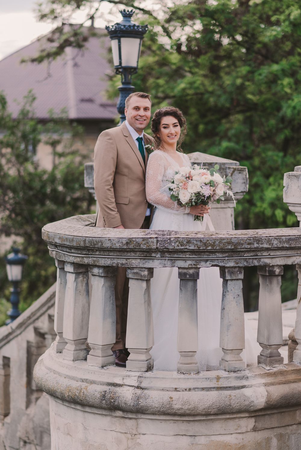 Newlyweds pose on historic stone balcony surrounded by greenery and vintage lampposts.