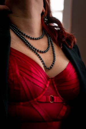 Black pearl necklace draped over red satin corset in dramatic lighting.