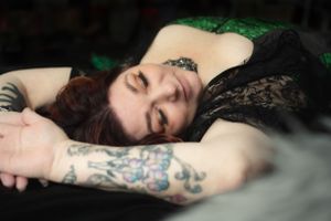 Person with tattoos reclining in black lace lingerie against dark background with green accent lighting.