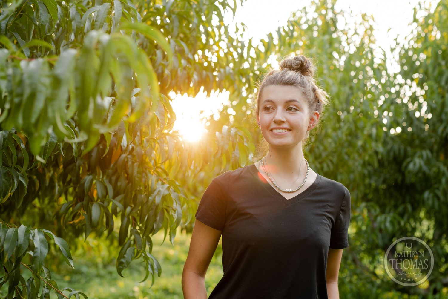 Natural portrait in black v-neck tee with sunset glowing through orchard trees.