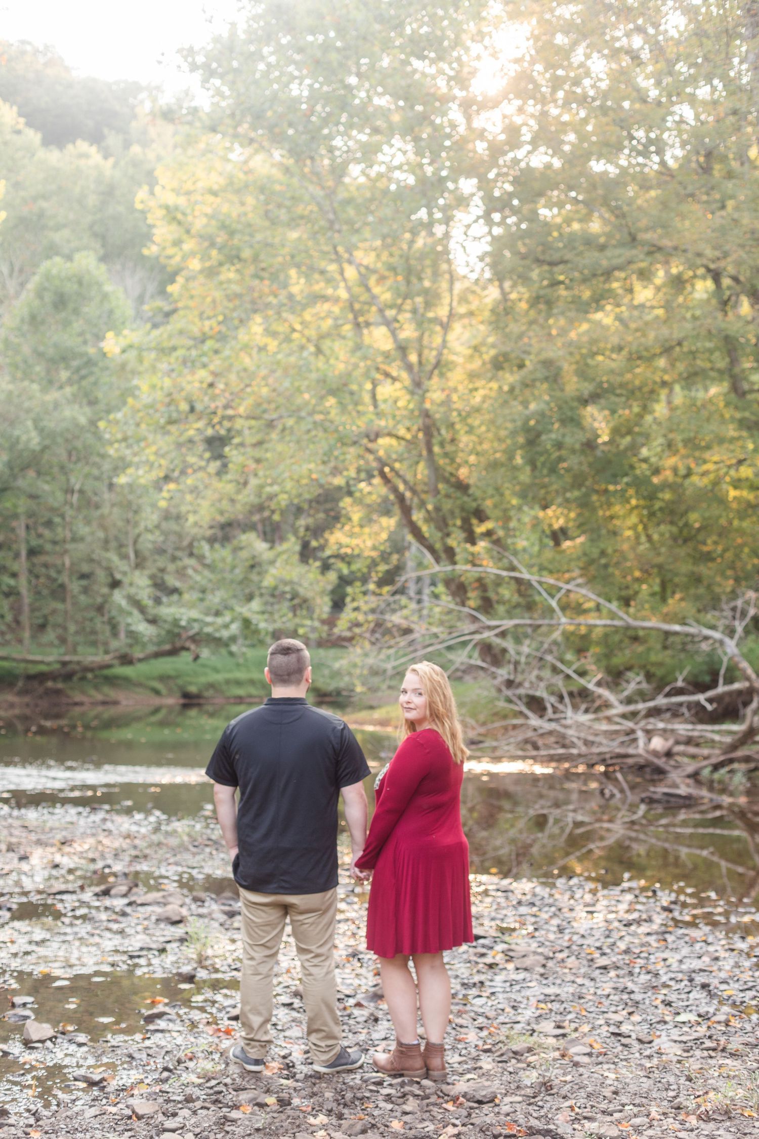 A couple walks along a rocky riverbank surrounded by autumn trees.