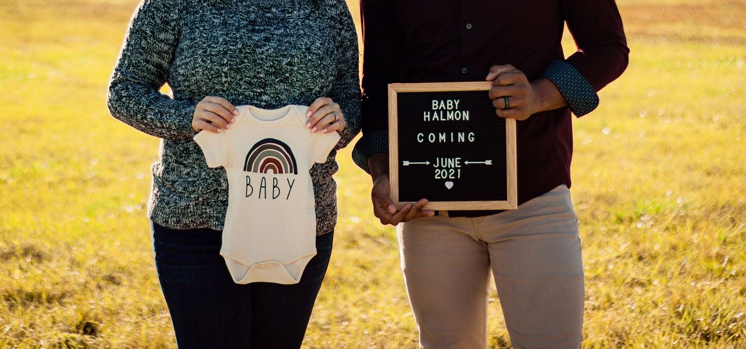 Couple stands in a sunny field holding a baby onesie and Baby Halmon Coming June 2021 sign.