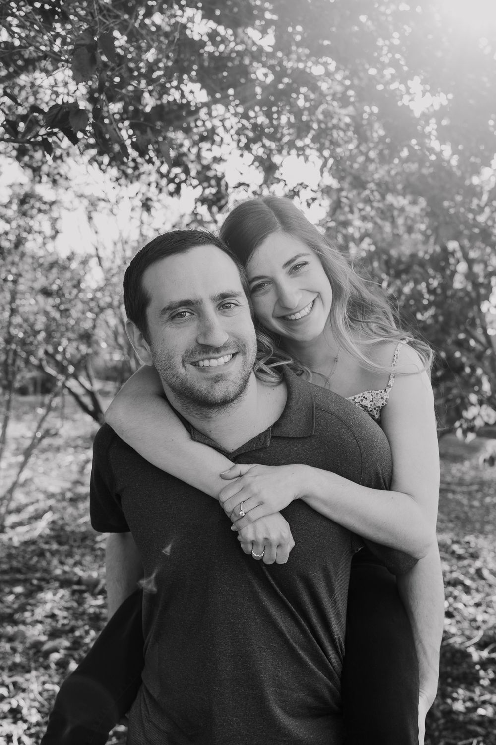 A black and white portrait of a couple sharing a piggyback ride outdoors.