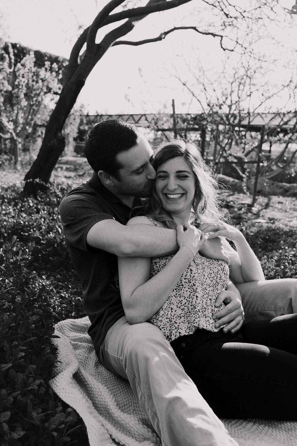 Black and white photo of couple cuddling on blanket with bridge in background.