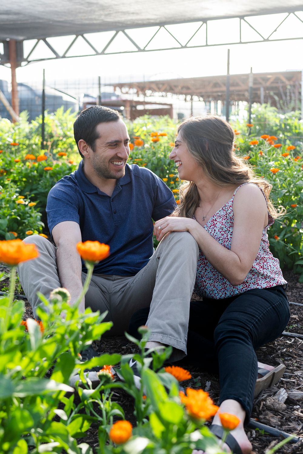 Couple sharing laughs while sitting among orange flowers in greenhouse garden.