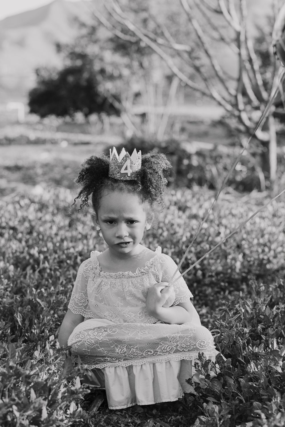 A young child in a white dress and crown poses in a garden setting, captured in black and white.