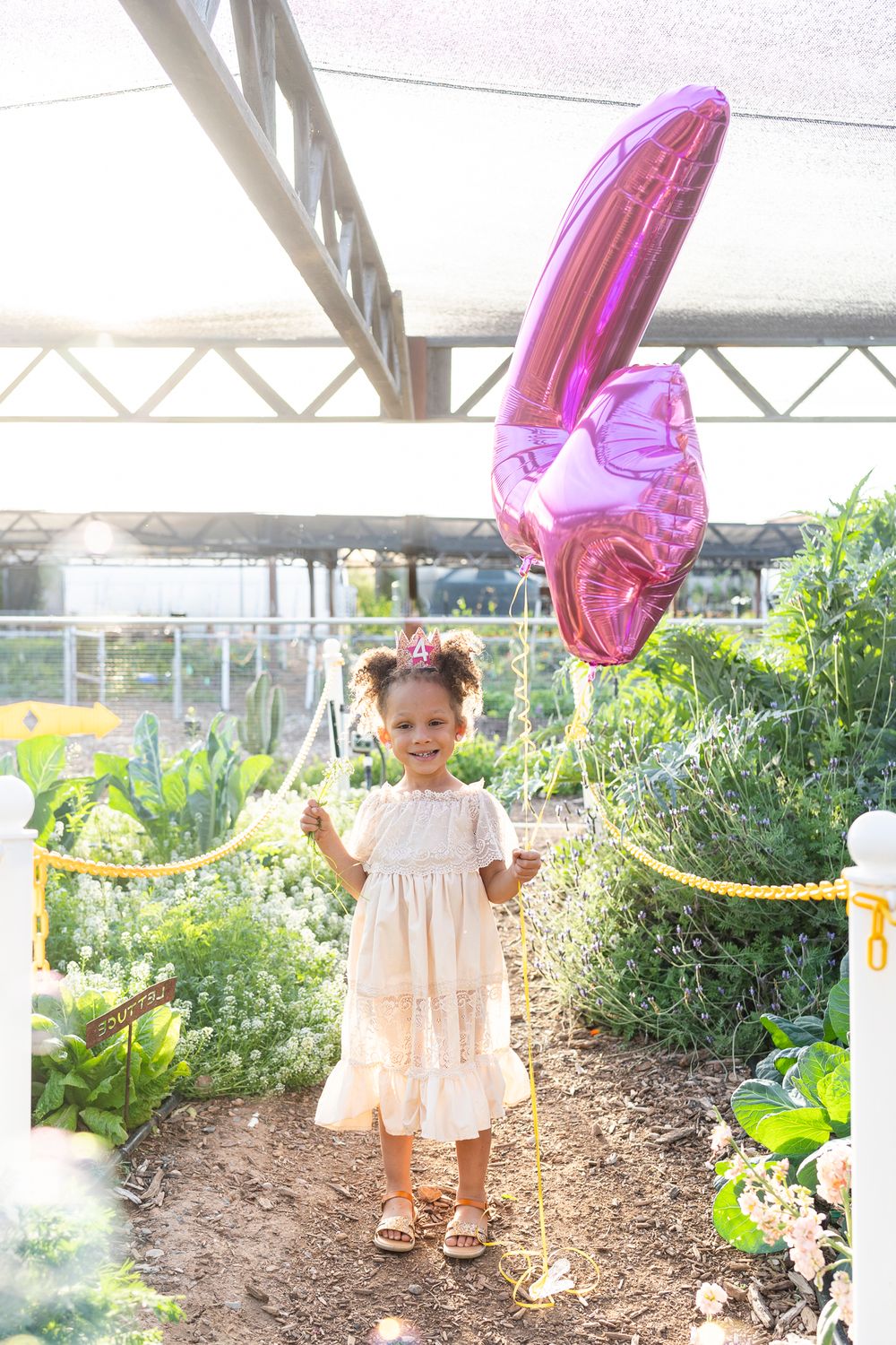 A child in a cream dress poses with a pink balloon in a sunny garden setting.