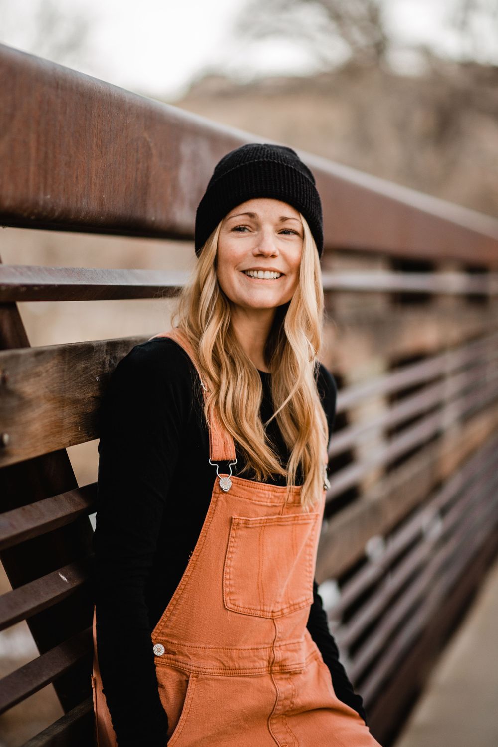 Smiling blonde woman in orange overalls and black beanie leaning against a wooden bridge railing in Golden Colorado.