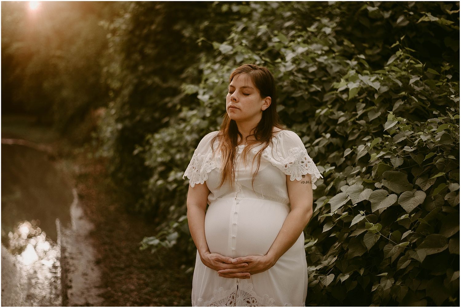 Moody maternity portrait with sunlight filtering through leaves as subject cradles belly.