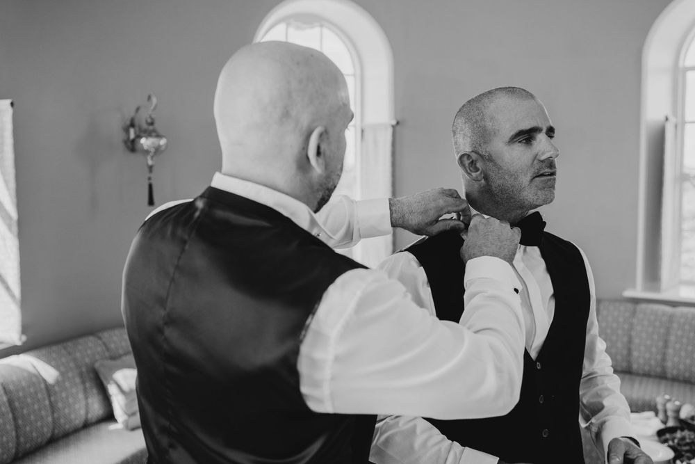 Two persons adjusting a formal tie before a wedding in a bright room with arched windows.