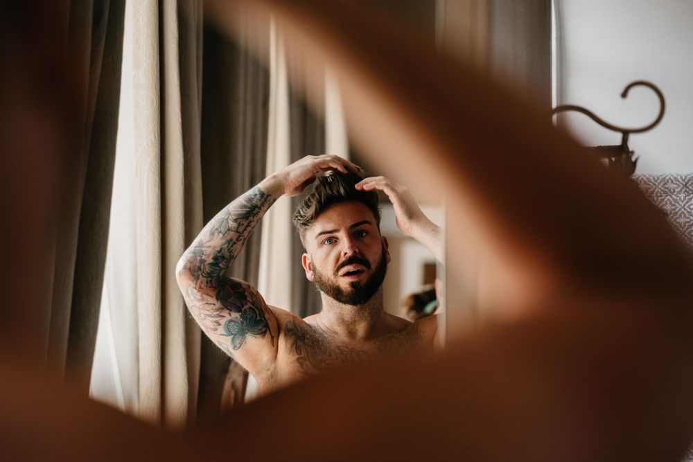 Mirror reflection shows a person with tattoos styling their hair in morning light.