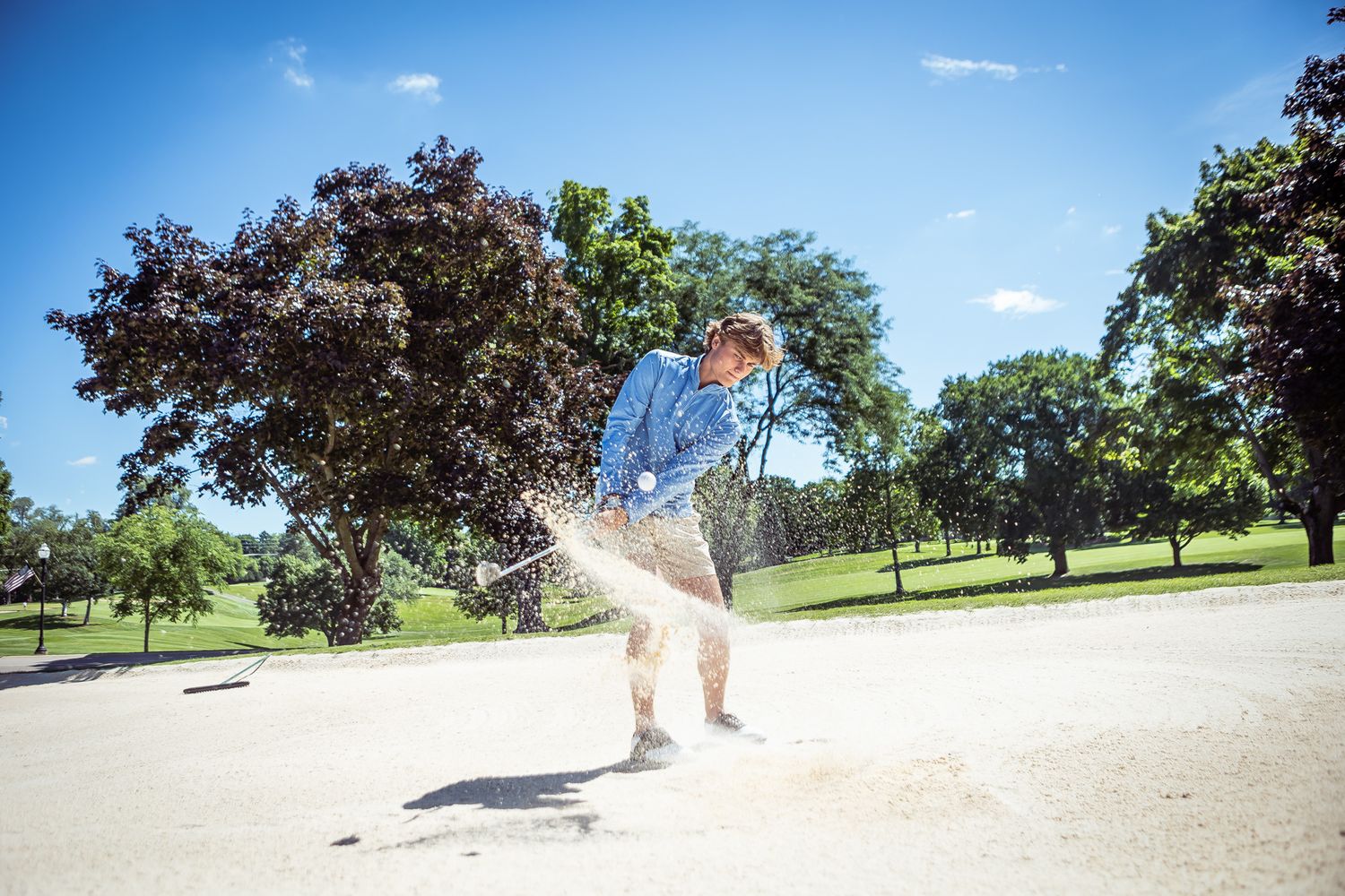A golf ball in mid-bounce on a paved trail surrounded by trees and grass on a sunny day.