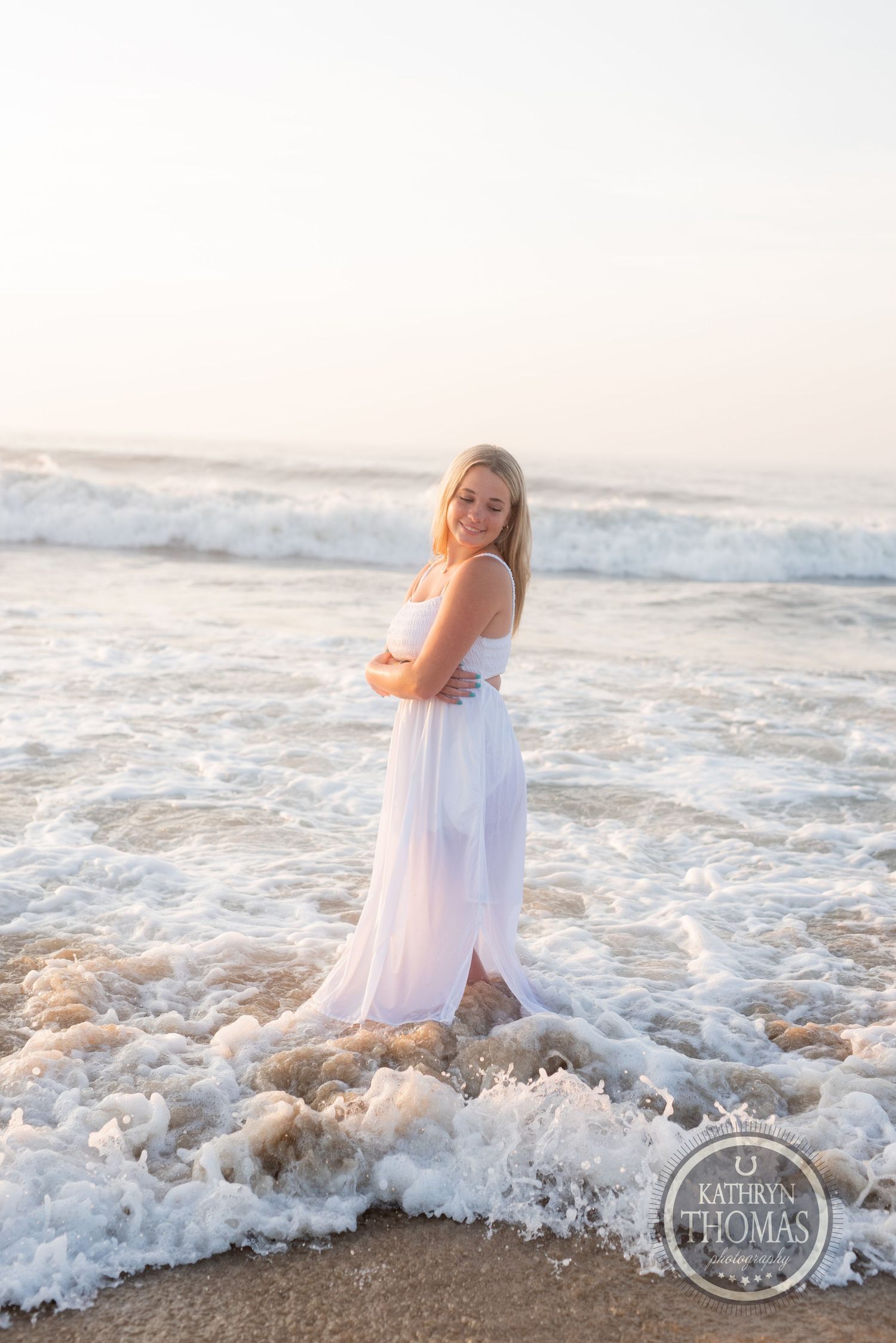 Person in white dress stands in ocean surf as waves crash around their feet at sunset.