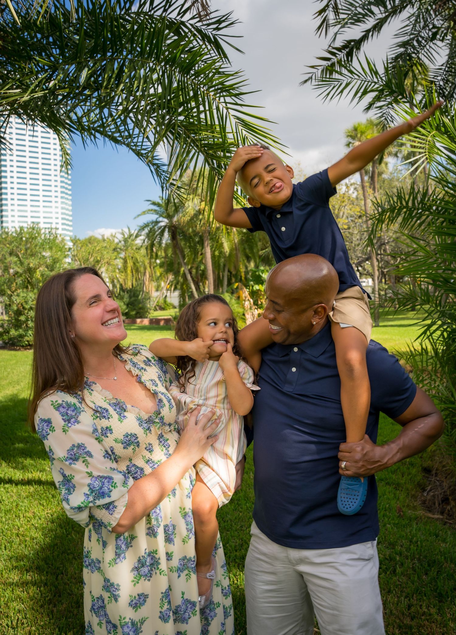 Happy family enjoying time together in a tropical park with palm trees and high-rise buildings.