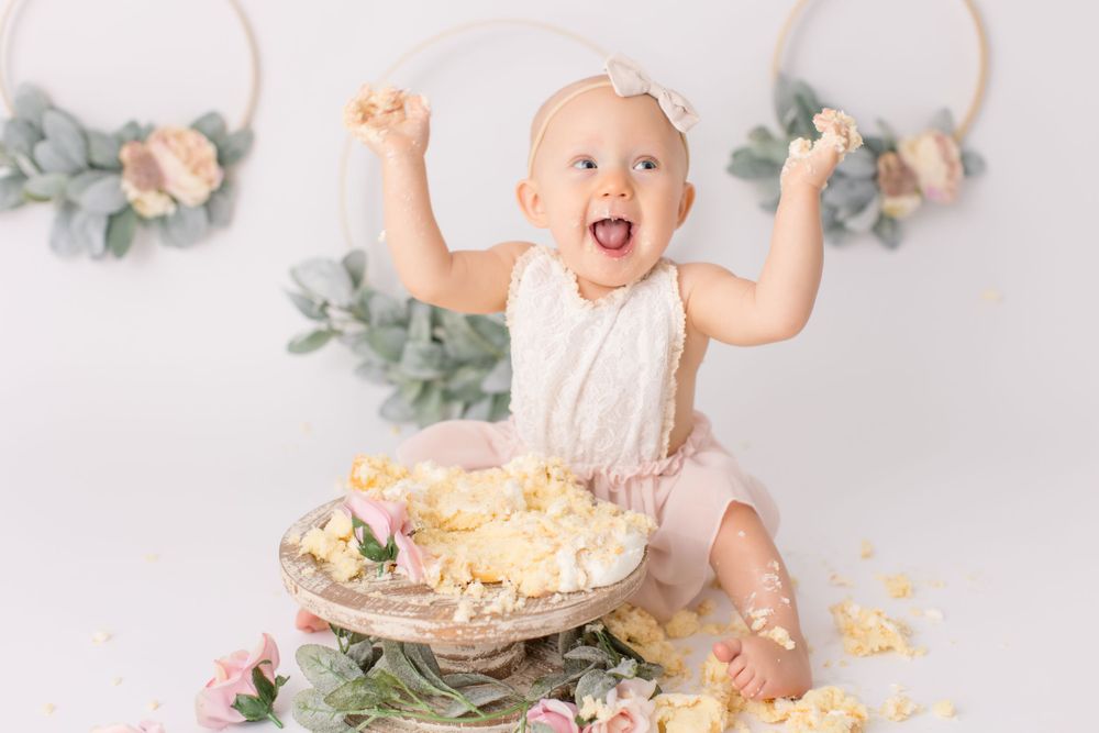 Excited baby throwing hands up during cake smash session with floral decorations.