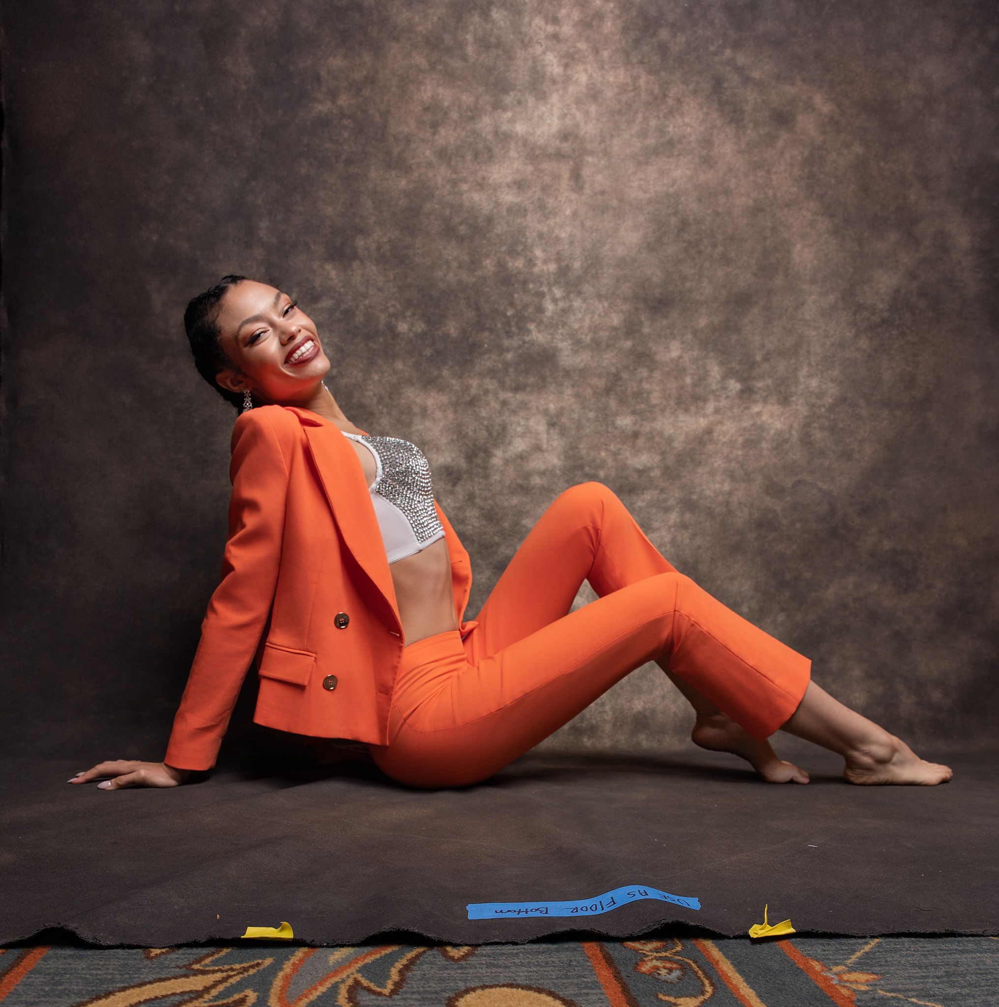 Woman in bright orange suit laughing while posing on floor against textured brown backdrop.